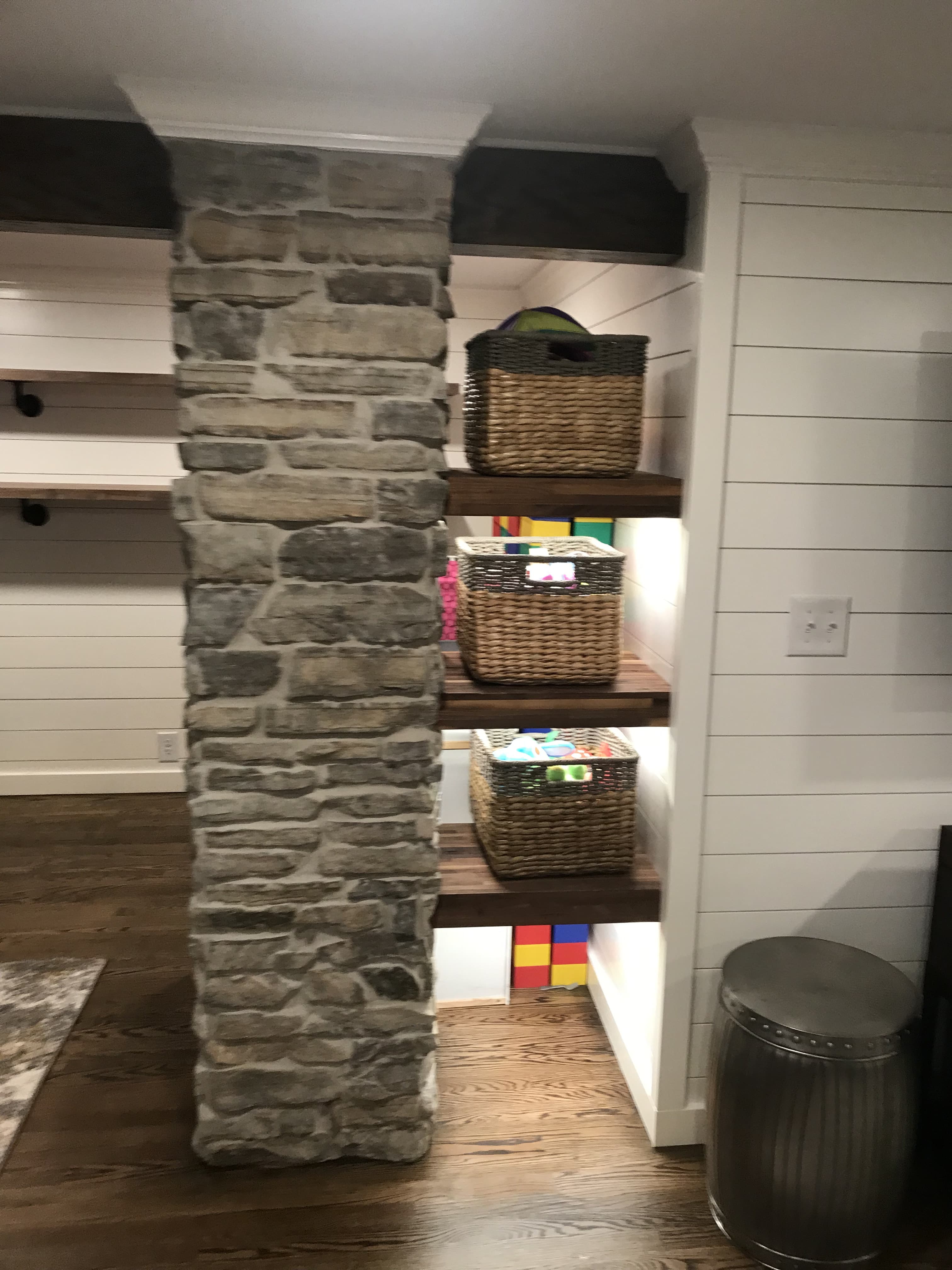 Grey stone pillar beside wooden shelves holding woven baskets against white shiplap walls.