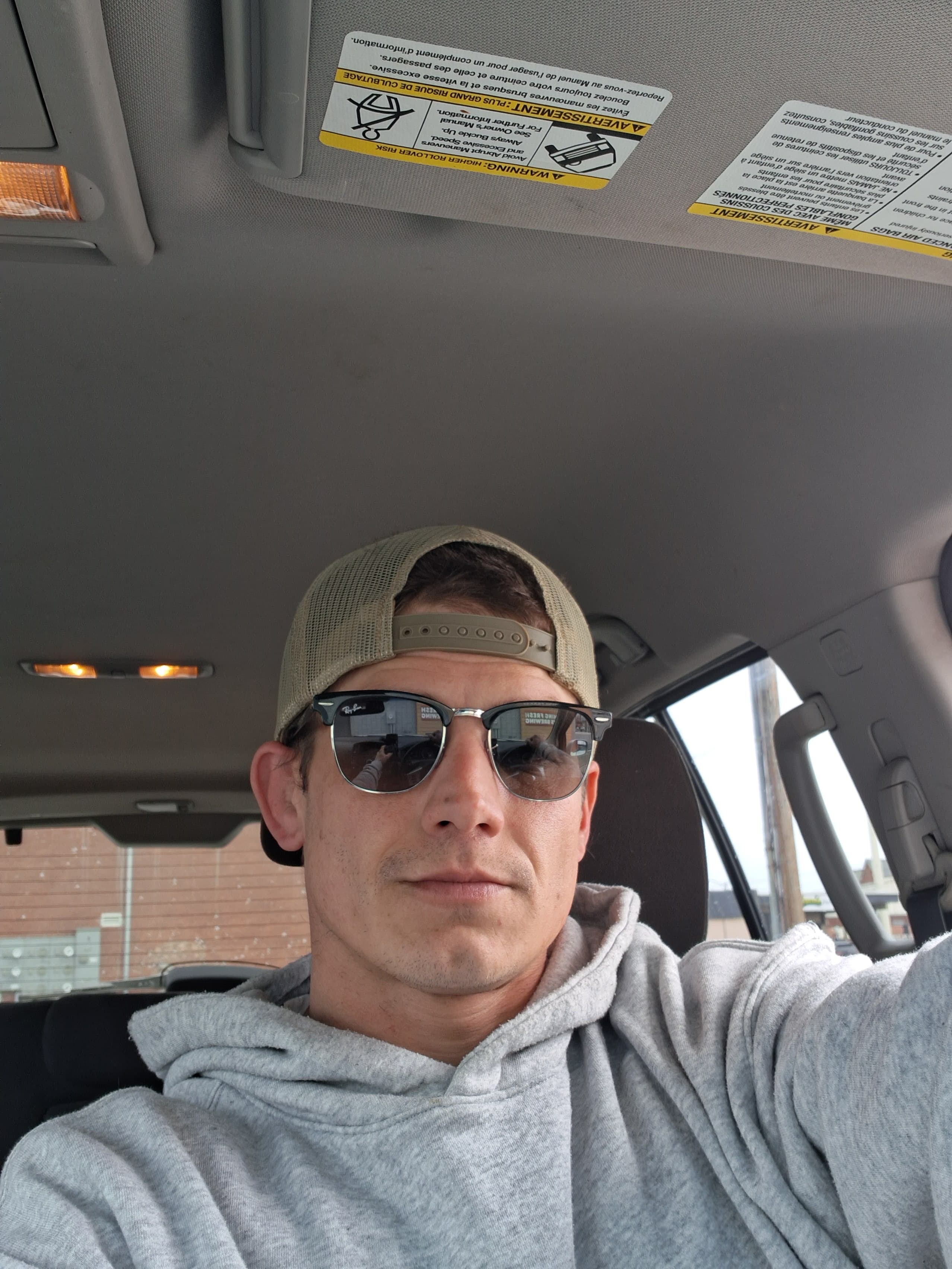 Man in a grey hoodie, sunglasses, and tan baseball cap sitting inside a car.